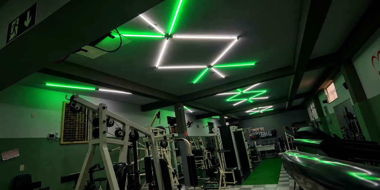 Academia com luminárias lineares de LED nas cores branca e verde formando figuras geométricas no teto