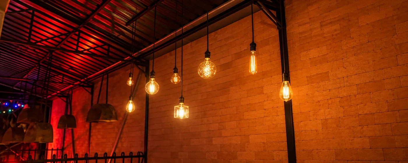 Lâmpadas pendentes de formatos variados acesas diante de uma parede de tijolos, sob estrutura metálica com luz vermelha